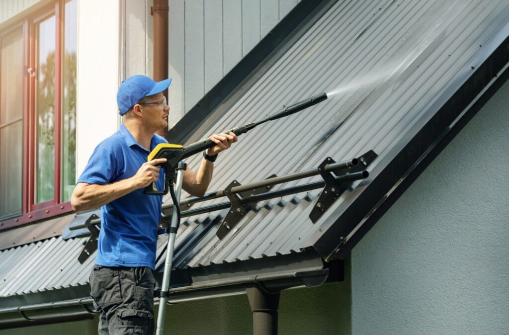 stock-photo-man-standing-on-ladder-and-cleaning-house-metal-roof-with-high-pressure-washer-1446080288-transformed-min
