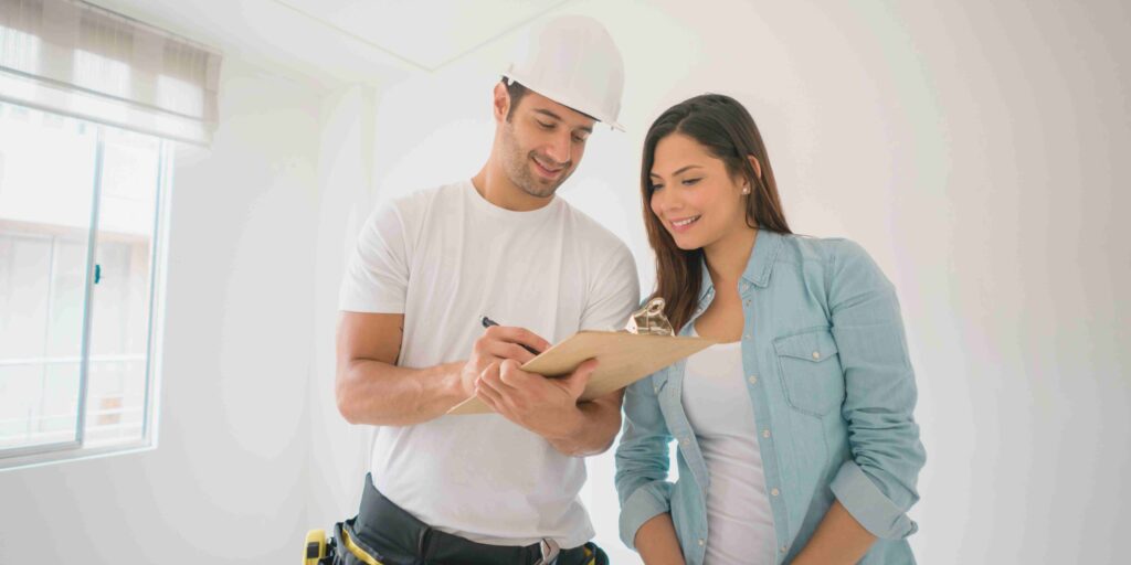 A man and woman in construction attire are reviewing plans together while holding a clipboard on a job site.