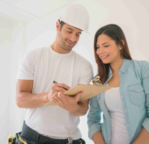 A man and woman in construction attire are reviewing plans together while holding a clipboard on a job site.